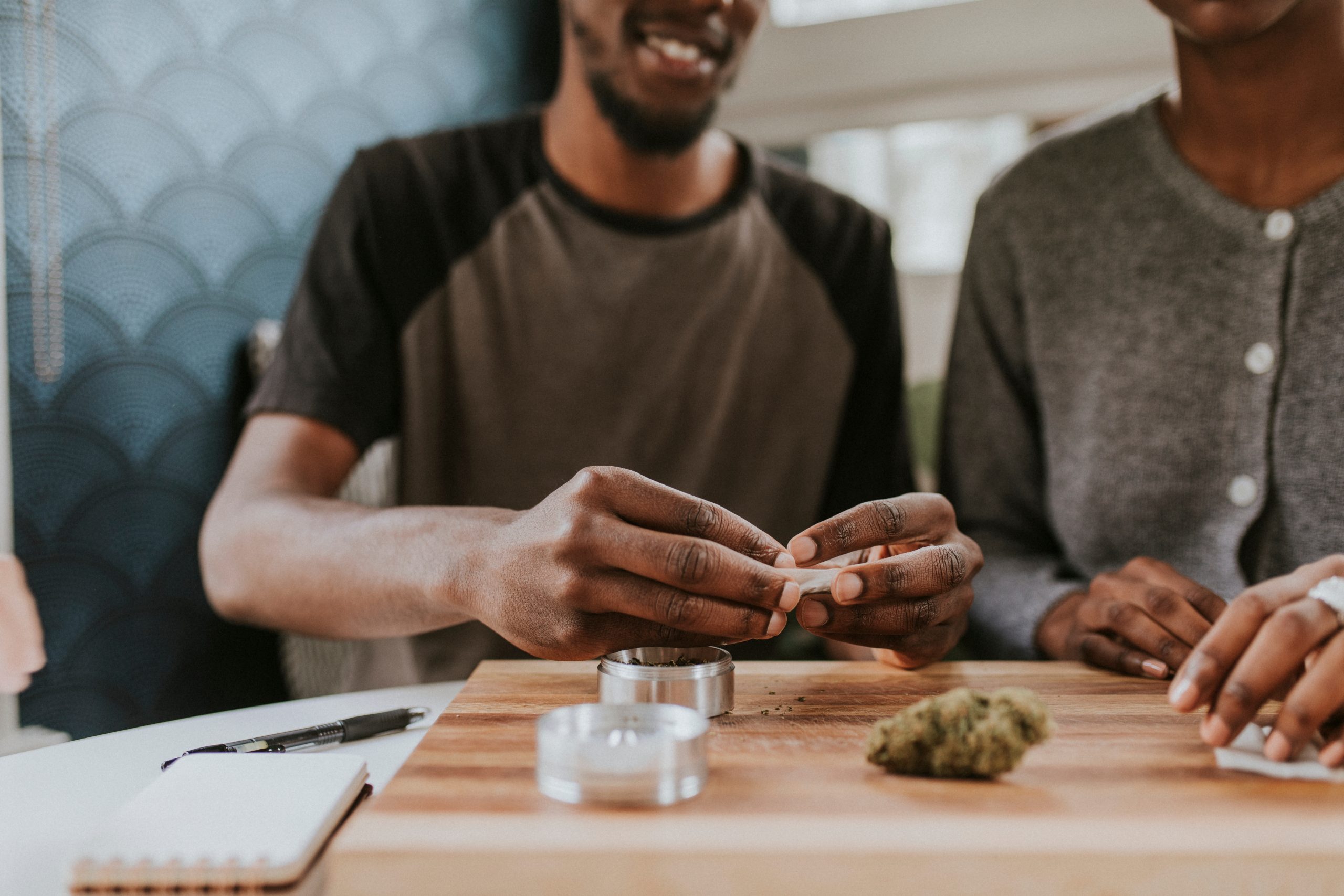 Two individuals rolling cannabis with a grinder, papers and cannabis nugget on the table.